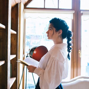 May include: A woman in a white blouse with puffy sleeves and a dark blue skirt stands near a wooden bookshelf, holding a book. A red lamp and a window with natural light are in the background.