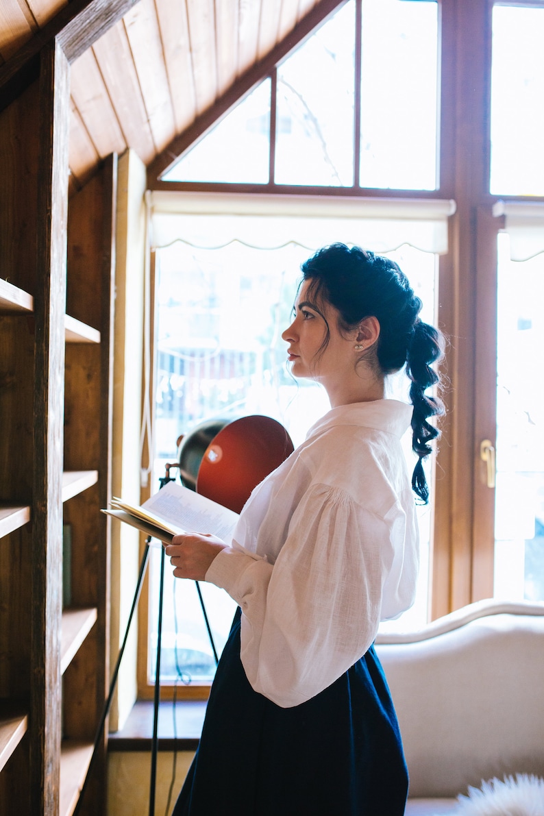 May include: A woman in a white blouse with puffy sleeves and a dark blue skirt stands near a wooden bookshelf, holding a book. A red lamp and a window with natural light are in the background.