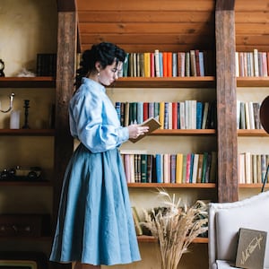 K&ouml;nnte beinhalten: Eine Frau in einem hellblauen Hemd und Rock steht in einem Raum mit B&uuml;cherregalen und liest ein Buch. Sie tr&auml;gt t&uuml;rkisfarbene Abs&auml;tze. Die B&uuml;cherregale sind mit B&uuml;chern gef&uuml;llt und auf dem Regal darunter steht eine Vase mit getrocknetem Weizen.