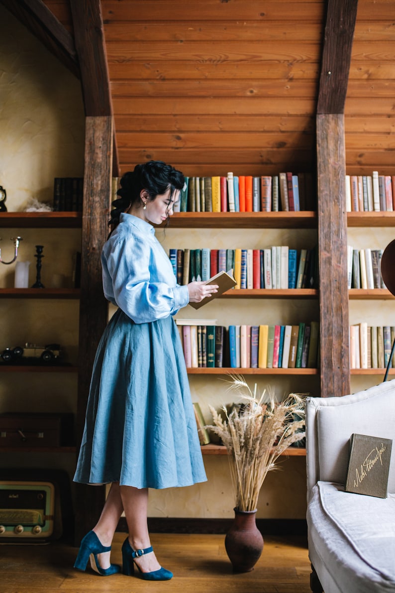 K&ouml;nnte beinhalten: Eine Frau in einem hellblauen Hemd und Rock steht in einem Raum mit B&uuml;cherregalen und liest ein Buch. Sie tr&auml;gt t&uuml;rkisfarbene Abs&auml;tze. Die B&uuml;cherregale sind mit B&uuml;chern gef&uuml;llt und auf dem Regal darunter steht eine Vase mit getrocknetem Weizen.