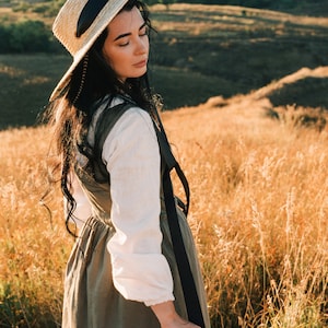 May include: A woman wearing a straw hat with a black ribbon and a long, flowing dress stands in a field of tall grass.