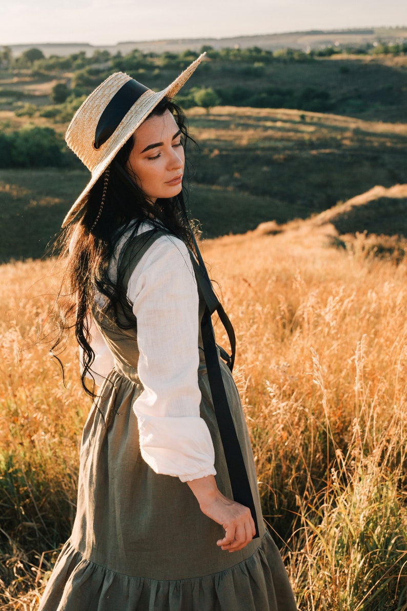 May include: A woman wearing a straw hat with a black ribbon and a long, flowing dress stands in a field of tall grass.
