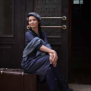 Puede incluir: Una mujer con un vestido azul y un gorro gris se sienta en una maleta vintage de madera marr&oacute;n. Lleva medias azules oscuras y botas con cordones. La mujer est&aacute; sentada frente a una puerta de madera con paneles de vidrio.