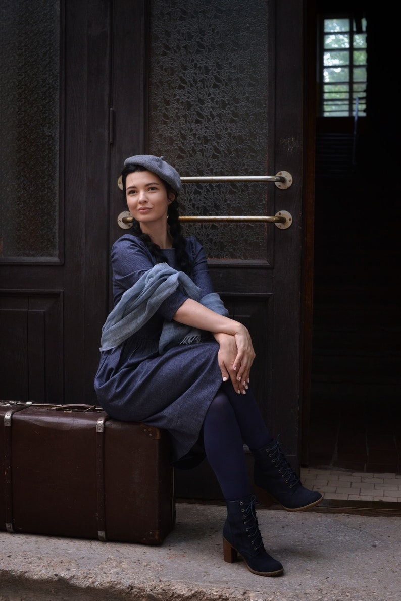 Puede incluir: Una mujer con un vestido azul y un gorro gris se sienta en una maleta vintage de madera marr&oacute;n. Lleva medias azules oscuras y botas con cordones. La mujer est&aacute; sentada frente a una puerta de madera con paneles de vidrio.