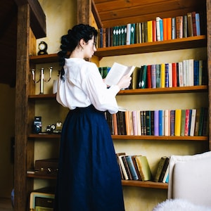 May include: A person wearing a white blouse and a long, navy blue skirt stands before a wooden bookshelf filled with books. The room has a vintage aesthetic, with a wooden ceiling and warm lighting. A vintage clock and camera are on a shelf.