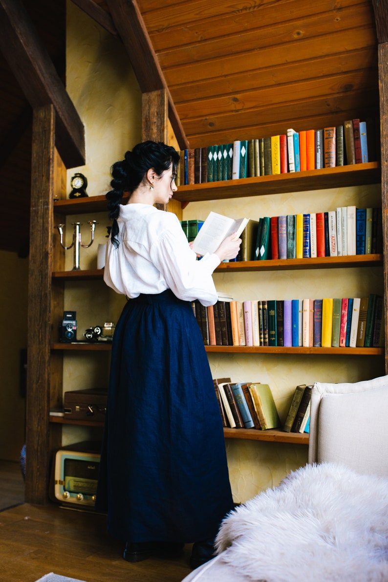 May include: A person wearing a white blouse and a long, navy blue skirt stands before a wooden bookshelf filled with books. The room has a vintage aesthetic, with a wooden ceiling and warm lighting. A vintage clock and camera are on a shelf.
