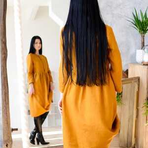 May include: A woman in a long, mustard yellow linen dress stands in front of a mirror, looking at her reflection. She is wearing black boots and has long, black hair.