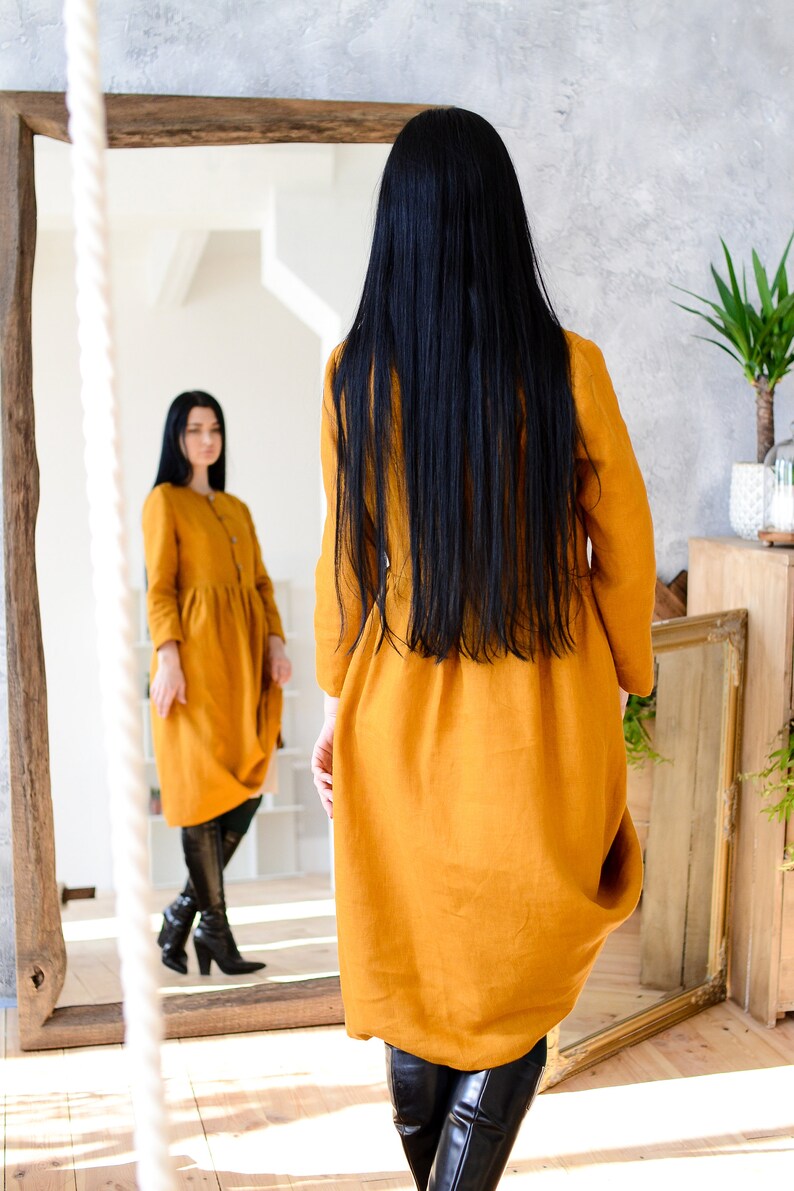 May include: A woman in a long, mustard yellow linen dress stands in front of a mirror, looking at her reflection. She is wearing black boots and has long, black hair.