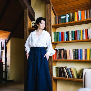 May include: A person wearing a white blouse and a long navy blue skirt, standing in a room with a wooden bookshelf. The skirt reaches the ankles. The room has wooden beams and a warm atmosphere. The bookshelf is filled with various books.