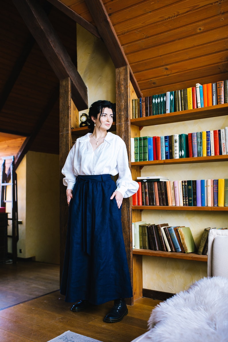 May include: A person wearing a white blouse and a long navy blue skirt, standing in a room with a wooden bookshelf. The skirt reaches the ankles. The room has wooden beams and a warm atmosphere. The bookshelf is filled with various books.