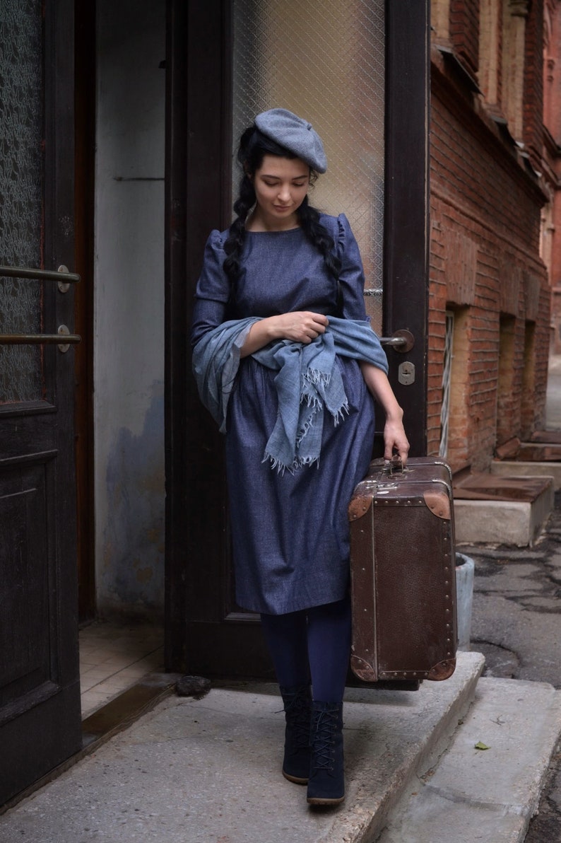 Puede incluir: Una mujer con un vestido de mezclilla azul y un gorro gris se encuentra frente a una puerta, sosteniendo una maleta vintage marr&oacute;n. Lleva medias azules y botas de cordones negras.