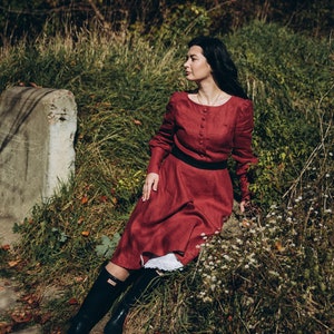May include: A woman wearing a long, burgundy dress with a black belt and black rain boots sits on a rock in a grassy area.