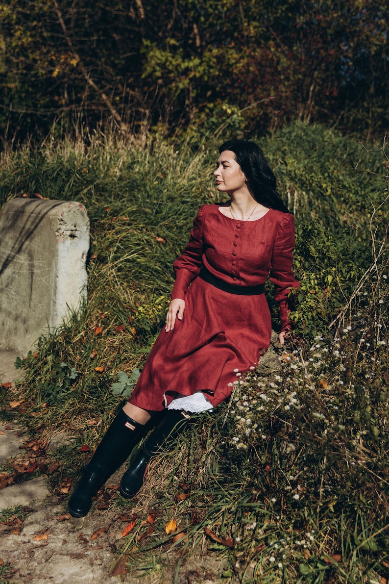 May include: A woman wearing a long, burgundy dress with a black belt and black rain boots sits on a rock in a grassy area.