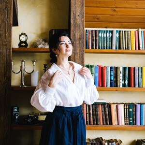 May include: A woman in a white blouse and a navy blue skirt stands before a bookshelf. The blouse has long sleeves and a collar. The skirt is a high-waisted, flowing style. A small clock and a candle holder are on a shelf.