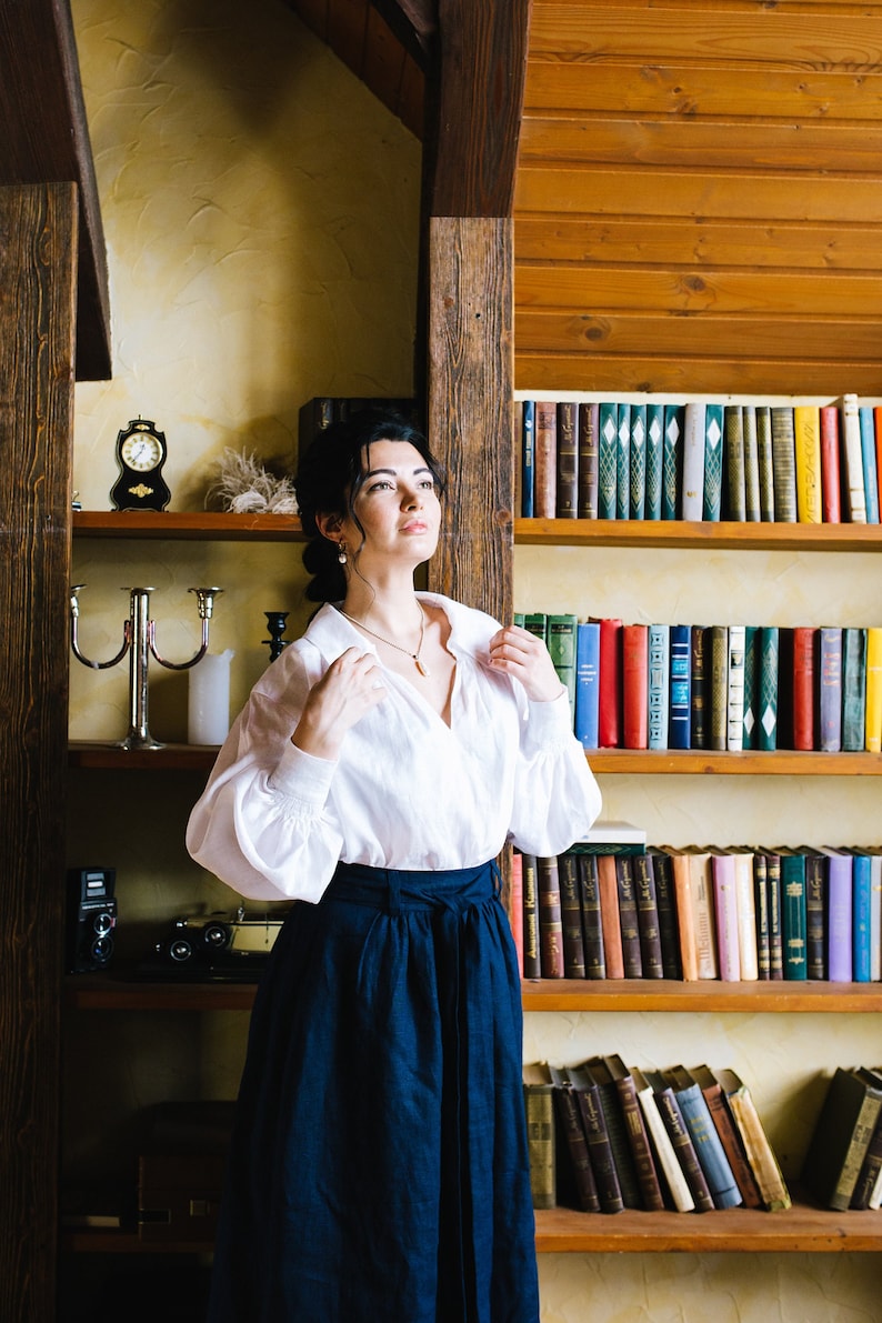 May include: A woman in a white blouse and a navy blue skirt stands before a bookshelf. The blouse has long sleeves and a collar. The skirt is a high-waisted, flowing style. A small clock and a candle holder are on a shelf.