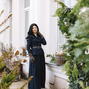 May include: A woman wearing a dark blue long-sleeved shirt and a matching long skirt. She is wearing a brown leather belt and brown boots. She is standing in front of a white building with a window and a green bush.