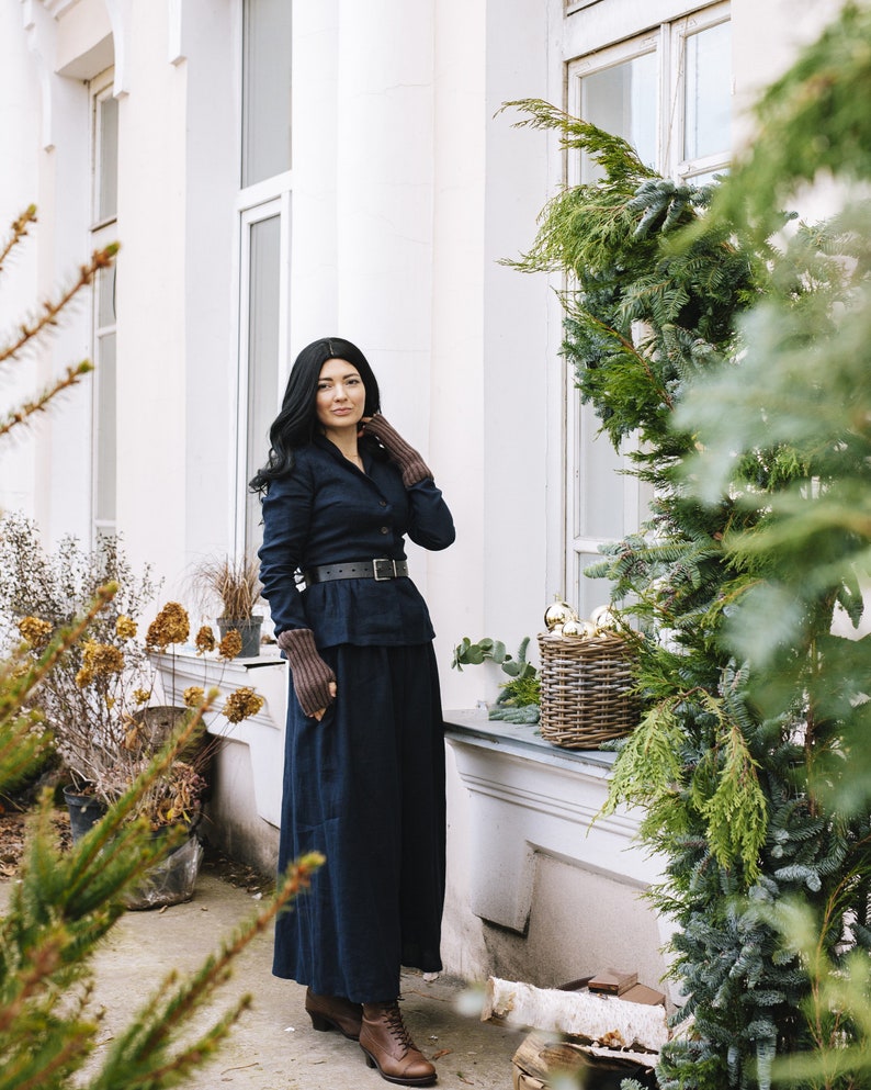 May include: A woman wearing a dark blue long-sleeved shirt and a matching long skirt. She is wearing a brown leather belt and brown boots. She is standing in front of a white building with a window and a green bush.