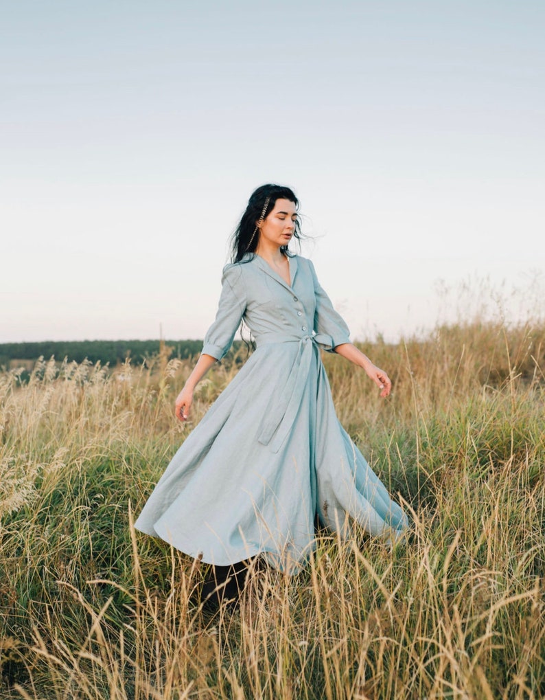 Puede incluir: Una mujer con un vestido azul claro con cintur&oacute;n se encuentra en un campo de hierba alta. El vestido tiene una falda larga y un cuello. La mujer mira hacia abajo y su cabello se mueve con el viento.