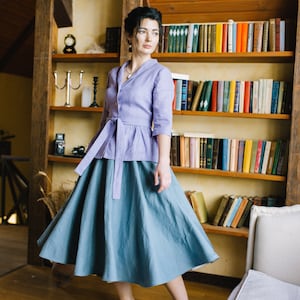 Op de afbeelding: Een vrouw draagt een lichtpaarse linnen blouse met een strik in de taille en een lange, zwierige blauwe rok. Ze staat in een kamer met een boekenkast achter haar.