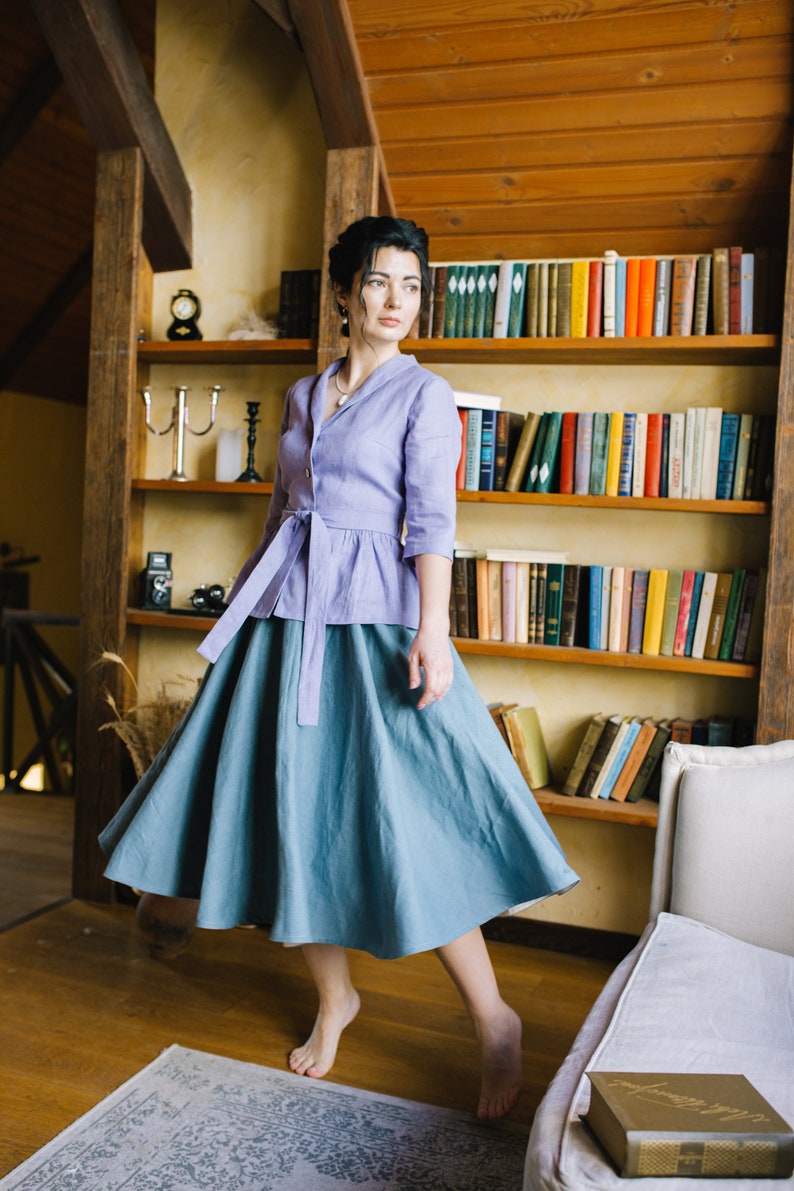 Op de afbeelding: Een vrouw draagt een lichtpaarse linnen blouse met een strik in de taille en een lange, zwierige blauwe rok. Ze staat in een kamer met een boekenkast achter haar.