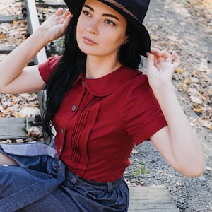 Puede incluir: Una mujer con un sombrero azul marino con una banda marr&oacute;n, una blusa roja de manga corta con la parte delantera plisada y una falda vaquera. La mujer est&aacute; sentada en las v&iacute;as del tren al aire libre.