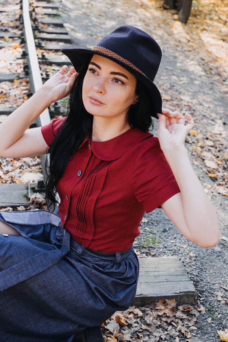 Puede incluir: Una mujer con un sombrero azul marino con una banda marr&oacute;n, una blusa roja de manga corta con la parte delantera plisada y una falda vaquera. La mujer est&aacute; sentada en las v&iacute;as del tren al aire libre.
