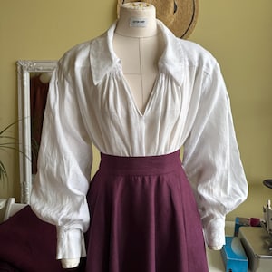 May include: A white linen blouse with a deep V-neck and voluminous sleeves is paired with a high-waisted, burgundy skirt. The outfit is displayed on a mannequin, suggesting a historical or theatrical costume. A straw hat hangs in the background.