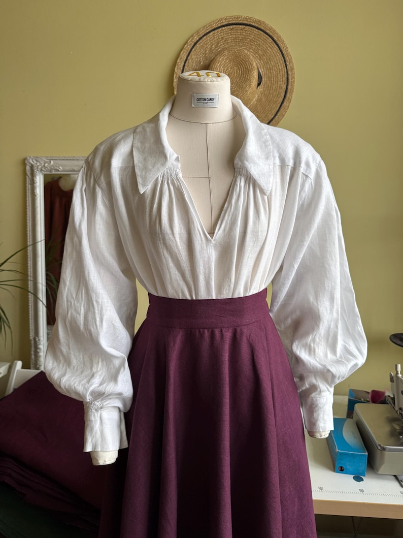 May include: A white linen blouse with a deep V-neck and voluminous sleeves is paired with a high-waisted, burgundy skirt. The outfit is displayed on a mannequin, suggesting a historical or theatrical costume. A straw hat hangs in the background.