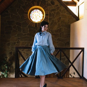K&ouml;nnte beinhalten: Eine Frau in einem hellblauen Hemd mit Kn&ouml;pfen und einem hellblauen Rock steht in einem Raum mit einer Holzdecke und einer Steinwand. Sie tr&auml;gt dunkelblaue Schuhe und ihr Rock weht im Wind.
