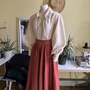 May include: A cream blouse with large sleeves and a wide collar, combined with a rust-coloured skirt. A straw hat hangs on the wall behind the mannequin. The outfit is displayed on a mannequin in a workshop setting.