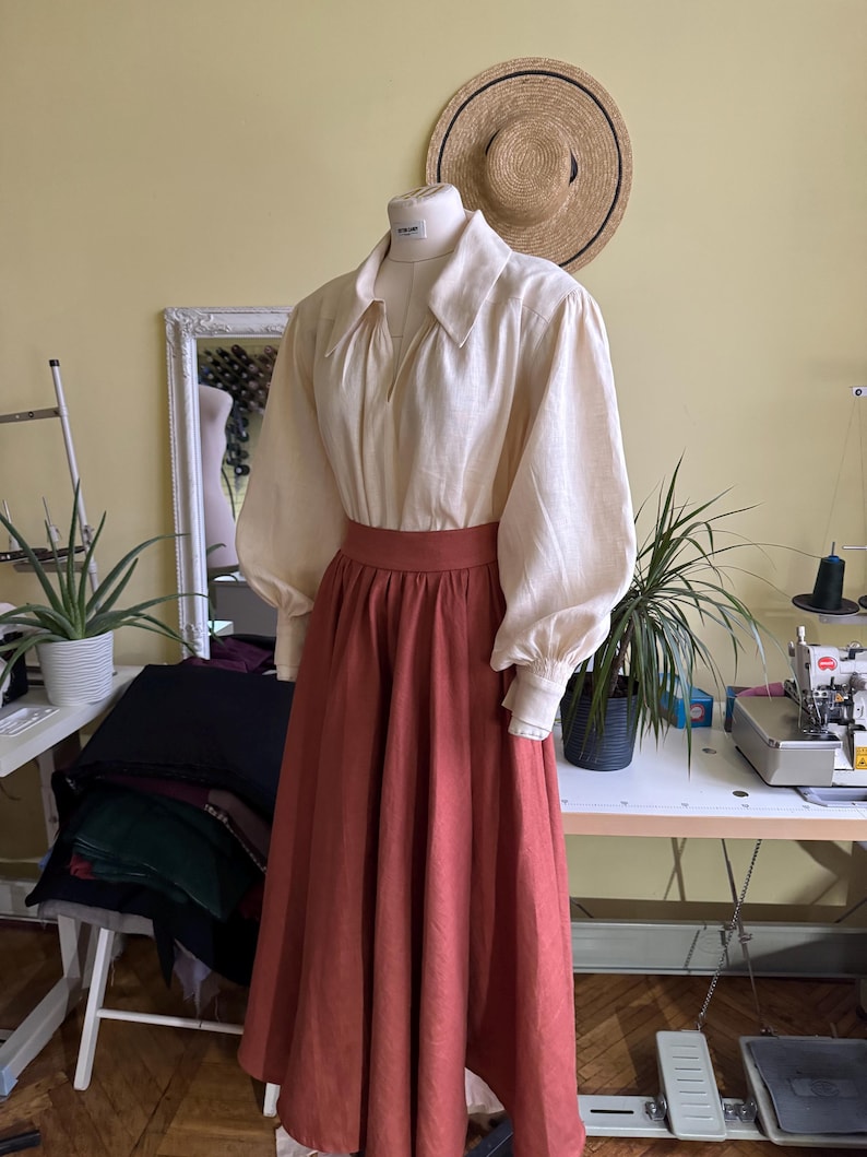 May include: A cream blouse with large sleeves and a wide collar, combined with a rust-coloured skirt. A straw hat hangs on the wall behind the mannequin. The outfit is displayed on a mannequin in a workshop setting.