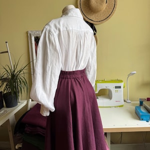 May include: A white blouse with long sleeves and a burgundy skirt are displayed on a mannequin. A straw hat hangs on the wall behind the mannequin. A sewing machine and other sewing supplies are on a table.