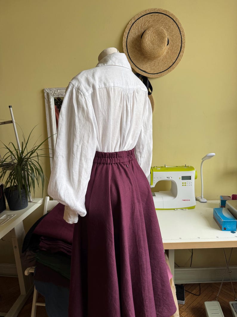May include: A white blouse with long sleeves and a burgundy skirt are displayed on a mannequin. A straw hat hangs on the wall behind the mannequin. A sewing machine and other sewing supplies are on a table.