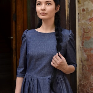 Puede incluir: Una mujer con el pelo largo y negro trenzado por la espalda lleva un vestido de mezclilla azul oscuro con cuello redondo y mangas abullonadas. Est&aacute; de pie frente a una puerta de madera y una pared de ladrillo.