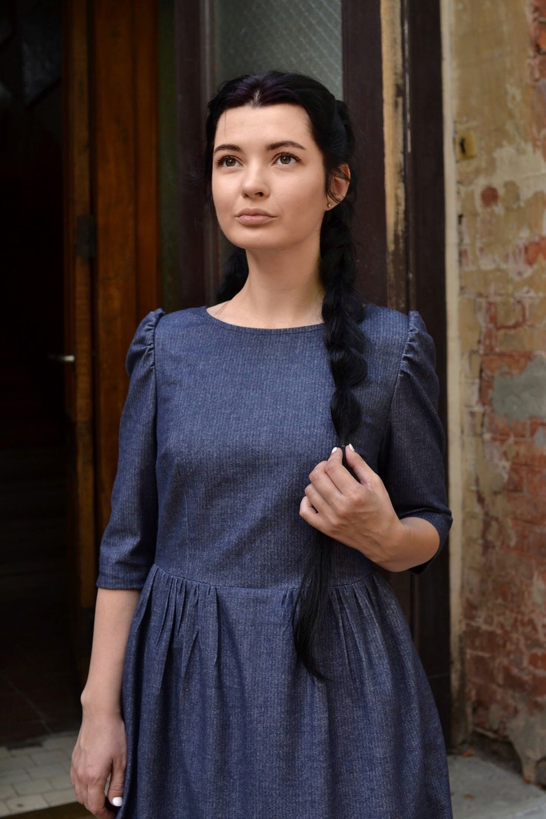 Puede incluir: Una mujer con el pelo largo y negro trenzado por la espalda lleva un vestido de mezclilla azul oscuro con cuello redondo y mangas abullonadas. Est&aacute; de pie frente a una puerta de madera y una pared de ladrillo.
