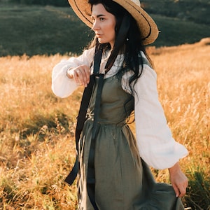 May include: A woman wearing a straw hat with a black ribbon tied around the crown, a white blouse, and a long green linen dress with a black ribbon tied around the waist. She is standing in a field of tall grass.