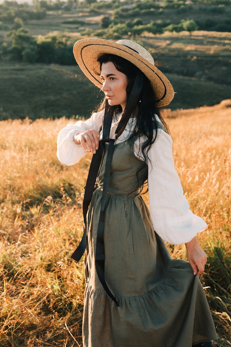 May include: A woman wearing a straw hat with a black ribbon tied around the crown, a white blouse, and a long green linen dress with a black ribbon tied around the waist. She is standing in a field of tall grass.