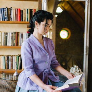 Op de afbeelding: Een vrouw in een lavendelkleurige linnen jurk met een blauwe rok zit in een kamer met boekenkasten en een spiegel. Ze leest een boek met een paarse kaft.