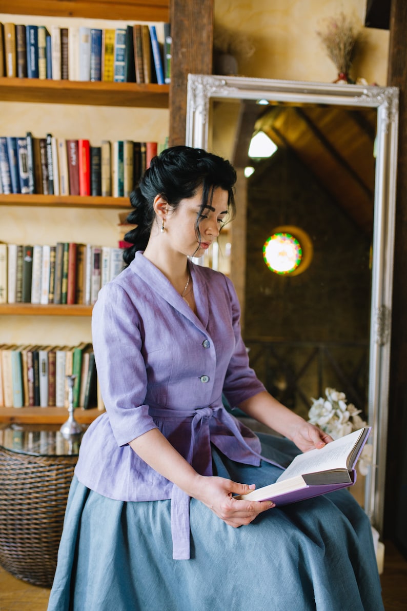 Op de afbeelding: Een vrouw in een lavendelkleurige linnen jurk met een blauwe rok zit in een kamer met boekenkasten en een spiegel. Ze leest een boek met een paarse kaft.