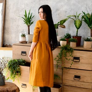 May include: A woman wearing a long, mustard yellow linen dress and black knee-high boots stands in front of a wooden dresser with open drawers. The dresser is decorated with potted plants.