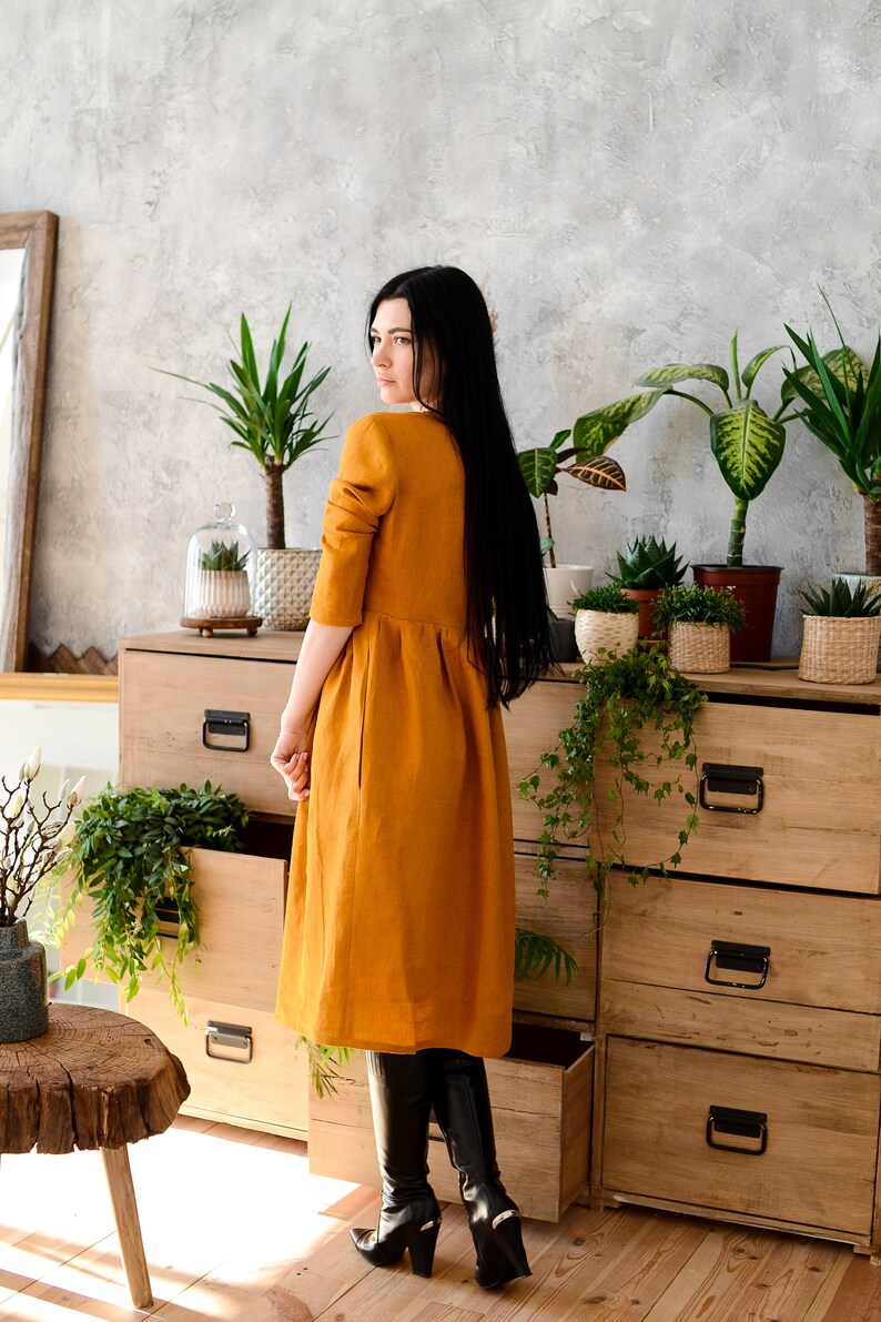 May include: A woman wearing a long, mustard yellow linen dress and black knee-high boots stands in front of a wooden dresser with open drawers. The dresser is decorated with potted plants.