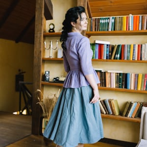 Op de afbeelding: Een vrouw draagt een lichtpaarse blouse met een gerimpelde zoom en een tealblauwe rok. Ze staat in een kamer met een boekenkast achter haar.