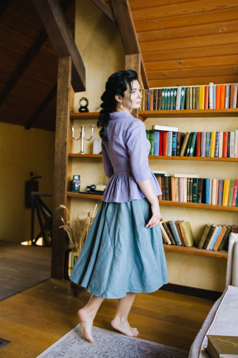 Op de afbeelding: Een vrouw draagt een lichtpaarse blouse met een gerimpelde zoom en een tealblauwe rok. Ze staat in een kamer met een boekenkast achter haar.