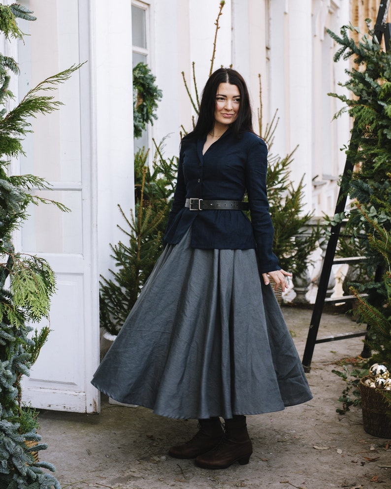 May include: A woman wearing a dark blue button-up shirt with a black belt and a long, gray skirt. She is standing in front of a white building with greenery.
