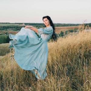 Puede incluir: Una mujer con un vestido azul claro se encuentra en un campo de hierba alta. El vestido ondea con el viento y la mujer mira a la c&aacute;mara.