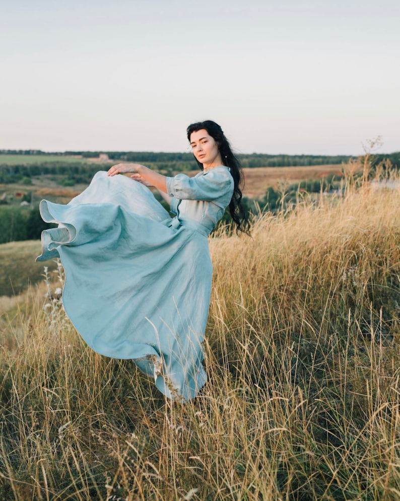 Puede incluir: Una mujer con un vestido azul claro se encuentra en un campo de hierba alta. El vestido ondea con el viento y la mujer mira a la c&aacute;mara.