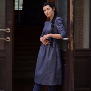 Puede incluir: Una mujer con un vestido de mezclilla azul oscuro y el pelo trenzado, de pie en una puerta. Lleva medias azules oscuras y botas negras con cordones.