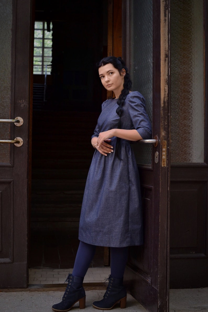 Puede incluir: Una mujer con un vestido de mezclilla azul oscuro y el pelo trenzado, de pie en una puerta. Lleva medias azules oscuras y botas negras con cordones.