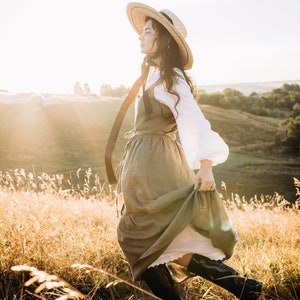 May include: A woman wearing a straw hat and a long, flowing green dress walks through a field of tall grass. The sun is shining brightly and the woman is smiling.