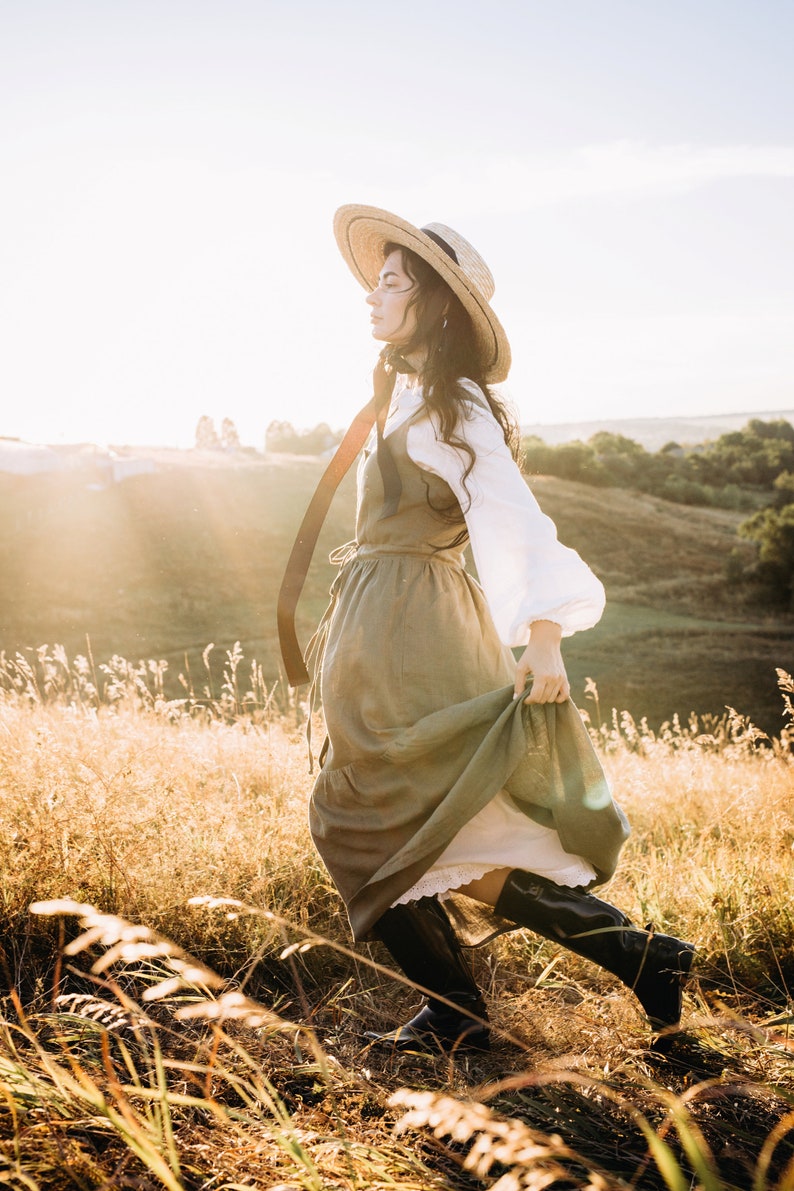 May include: A woman wearing a straw hat and a long, flowing green dress walks through a field of tall grass. The sun is shining brightly and the woman is smiling.